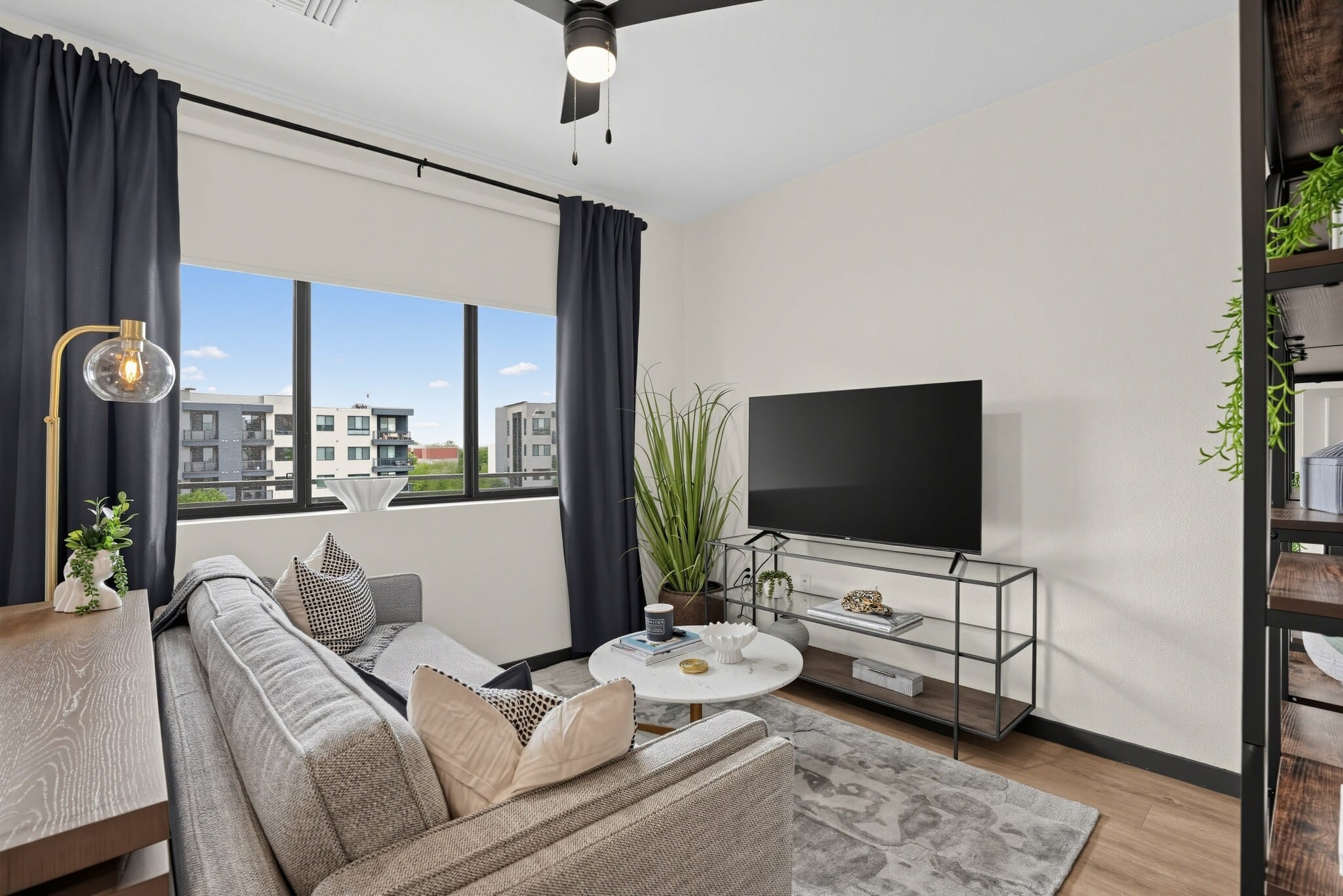 Modern living room with a gray sofa, round coffee table, TV on a stand, large window with black curtains, and potted plant. Shelving unit and ceiling fan also visible.