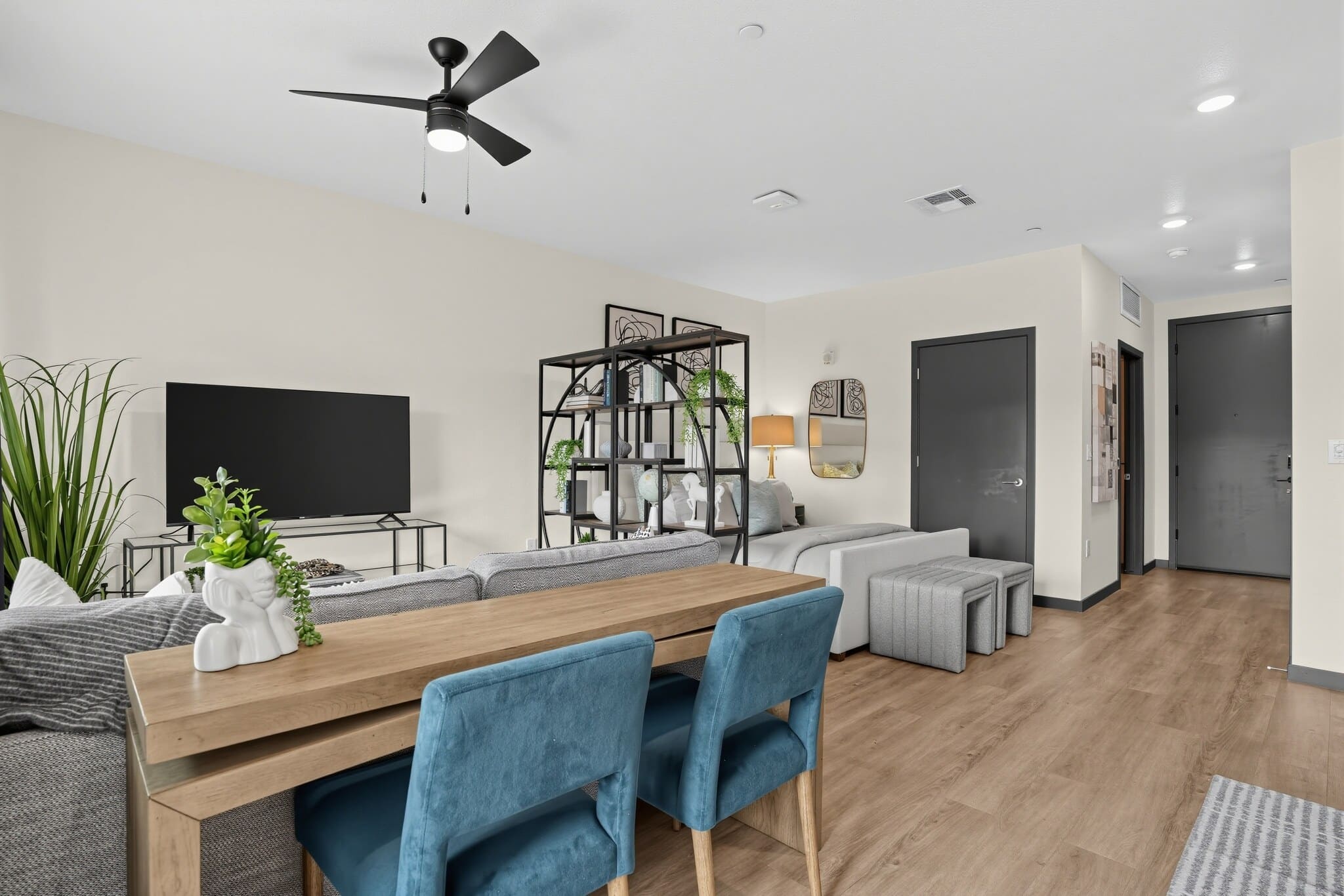 Modern living room with a wooden dining table, blue chairs, gray sofa, TV, metal bookshelf, and indoor plants on light wood flooring.