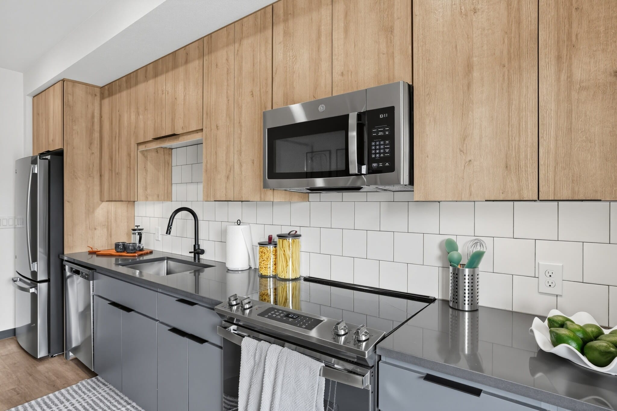 Modern kitchen with wood cabinets, gray countertops, stainless steel appliances, a bowl of limes, utensils in a holder, and a towel hanging on the oven door.