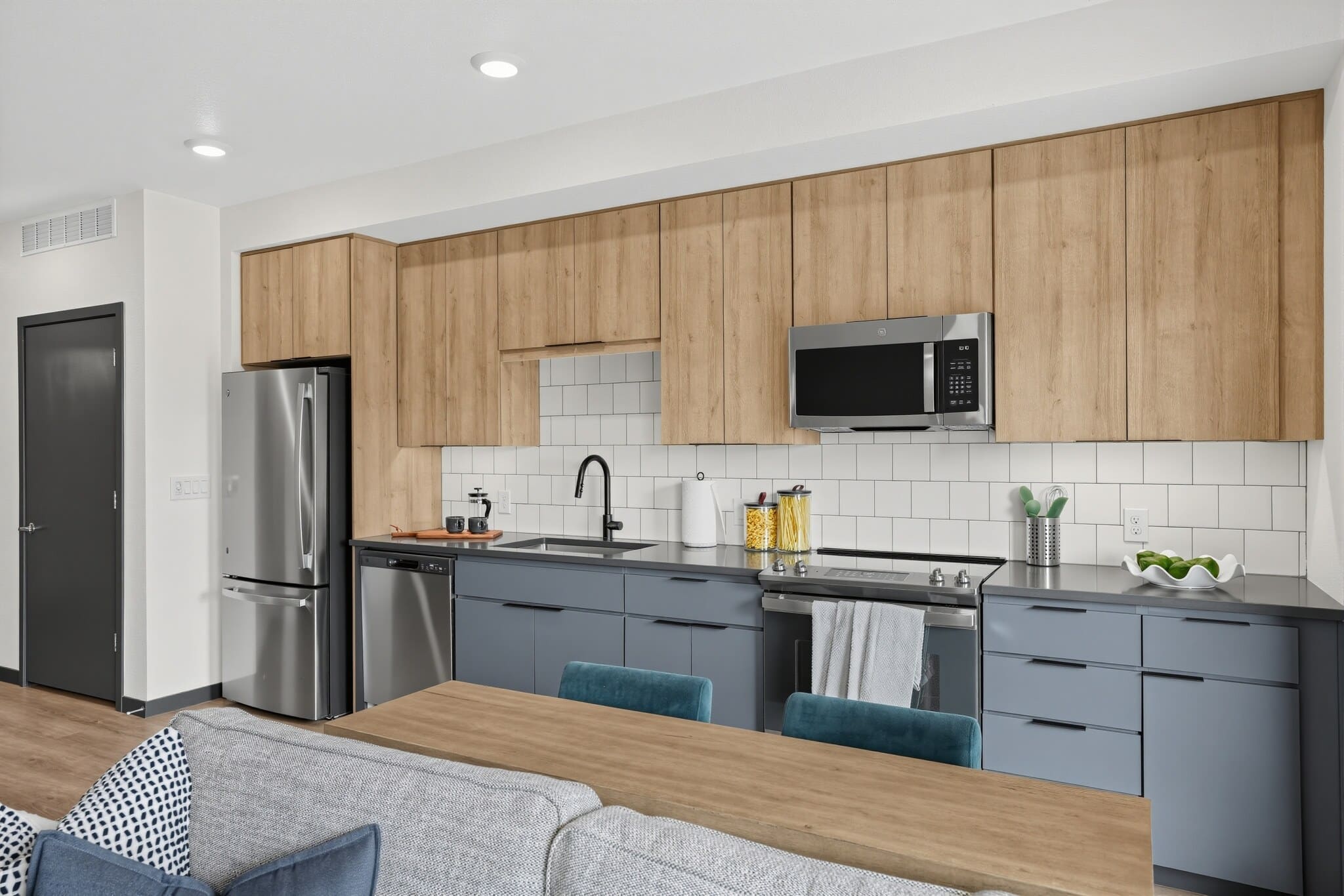 Modern kitchen with wooden upper cabinets, gray lower cabinets, stainless steel appliances, white tile backsplash, and a black faucet. Part of a living area is visible in the foreground.
