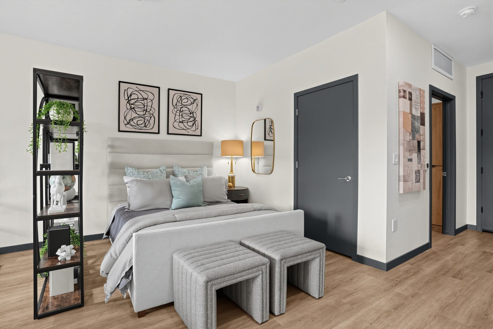 Modern bedroom with a gray bed, decorative pillows, two benches at the foot, a shelving unit with plants, abstract wall art, and wood flooring.