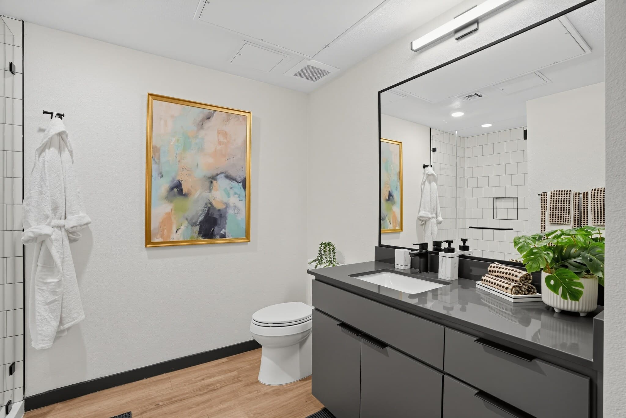 Modern bathroom with a black vanity, large mirror, toilet, abstract wall art, plants, and robes hanging near a glass shower. Light wood flooring contrasts with white walls.