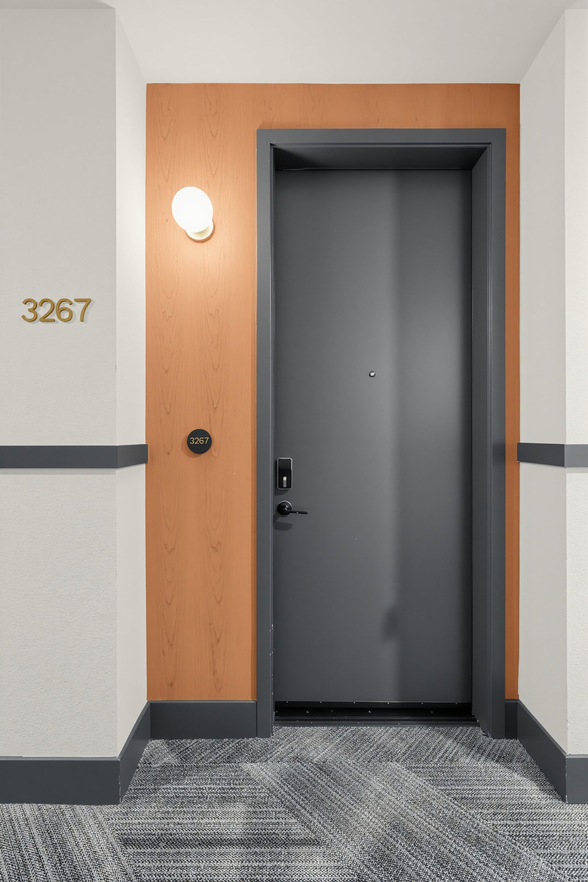 A dark gray door with a peephole and handle is set in a wood-paneled alcove. The number 3267 is displayed on the wall and door. A wall sconce is mounted to the left of the door.